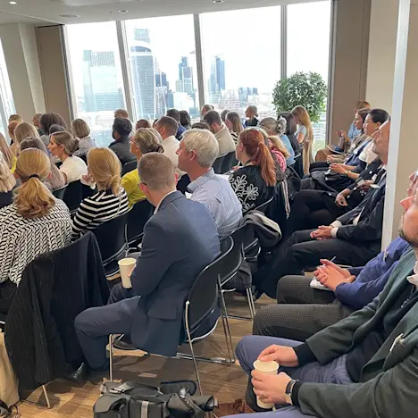 An engaged audience listens, as the City is seen in the background Tile image of An engaged audience listens, as the City is seen in the background