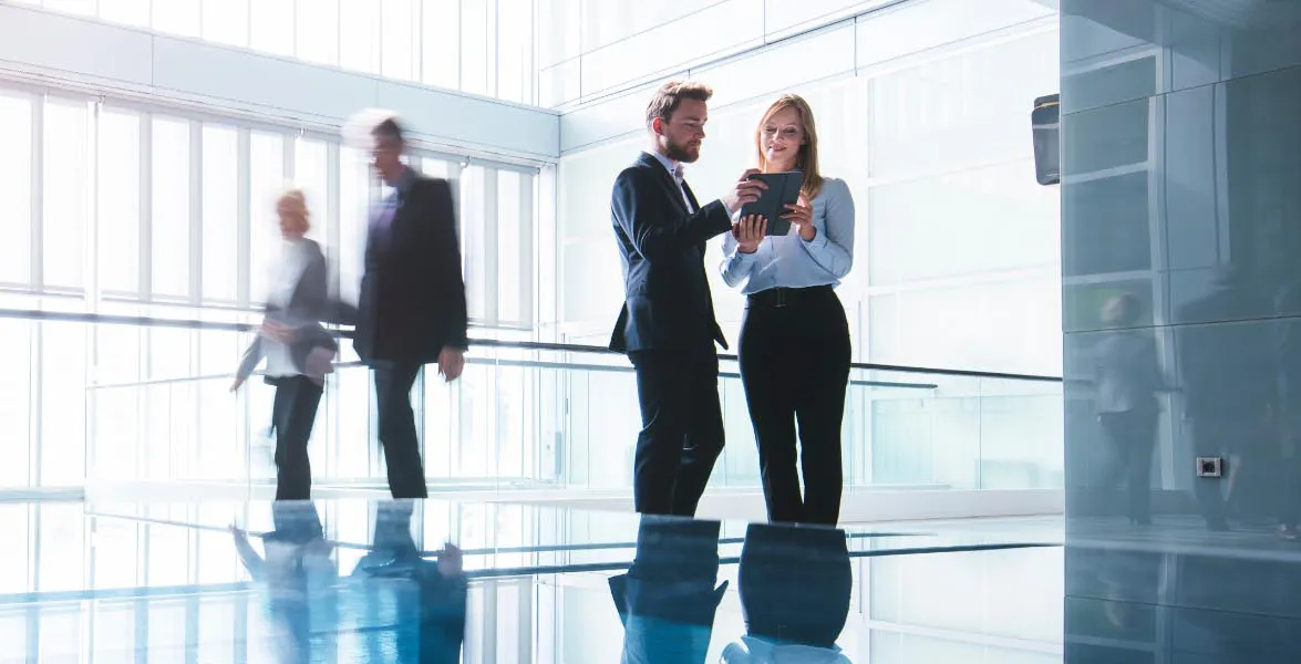 A businessman and a businesswoman looking at a tablet as people walk by